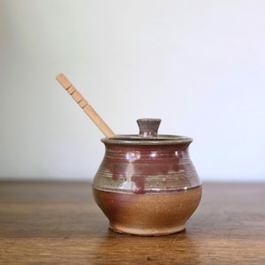May include: A ceramic honey pot with a rounded body, featuring a two-tone glaze of reddish-brown and earthy tan. The pot includes a matching lid with a small knob handle and a light-colored wooden honey dipper resting inside the opening.