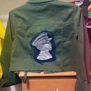 May include: Olive green jacket with a patch featuring a profile of a person in a military hat. The patch is white with black and blue accents. The jacket has a collar, long sleeves, and a button-up front.