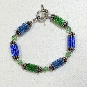 May include: A silver bracelet with alternating blue and green glass beads. The beads are faceted and have a striped pattern. The bracelet has a silver toggle clasp.