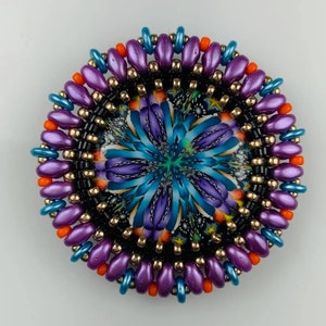 May include: A round beaded brooch with a purple, blue, and orange color scheme. The center of the brooch features a colorful, abstract design.