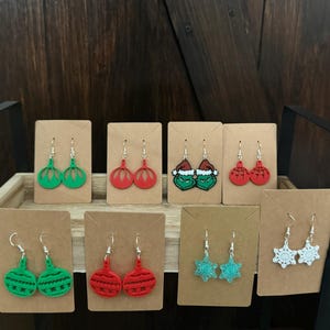 May include: Assortment of Christmas-themed earrings displayed on cards. The earrings feature various designs, including red and green ornaments, a Grinch character, and snowflakes.