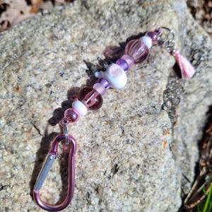 Pode incluir: Um porta-chaves de contas rosa e roxas com uma pequena figura de vidro branca e rosa. O porta-chaves inclui um mosquetão rosa e uma pequena borla rosa. As contas são de várias formas e tamanhos, incluindo redondas e oblongas.