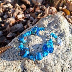 Op de afbeelding: Een blauwe glazen kralen armband met zilveren accenten. De kralen zijn gedraaid met witte en blauwe patronen. De armband ligt op een grijze steen.