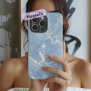 May include: A light blue smartphone case featuring a white marble-like pattern with subtle silver accents. The case has a rectangular cutout for the camera lens array and includes a pink label with the text Magsafe printed in black.