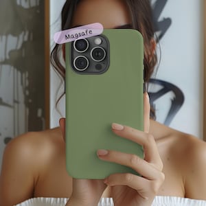 May include: Olive green phone case with a MagSafe label. The case is covering a smartphone with multiple camera lenses. The case is held up in front of a person.