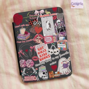 Puede incluir: Una funda negra para lector electrónico con un collage de pegatinas que incluye un esqueleto, un fantasma leyendo un libro, cartas del tarot, auriculares, dados y el texto "Hot Girls Read Books" y "Morally Grey is my favorite color."