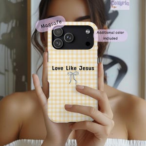 May include: A yellow and white gingham patterned phone case featuring the black text Love Like Jesus and a small line-art bow graphic. The case includes a black camera module cutout and labels indicating Magsafe and additional colour included.