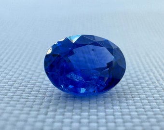 May include: A vibrant, oval-cut blue gemstone featuring a faceted surface that reflects light. The stone displays a deep, saturated royal blue color with visible internal inclusions and sharp, geometric edges across its polished top surface.