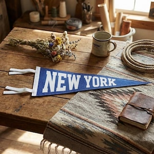 Puede incluir: Un banderín azul con las palabras "NEW YORK" en letras blancas descansa sobre una mesa de madera. Un pequeño ramo de flores secas, una taza, una bobina de cuerda y un diario encuadernado en cuero también están sobre la mesa. El banderín tiene lazos blancos.
