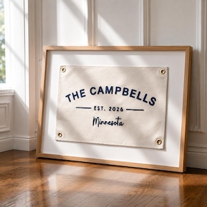 Op de afbeelding: Een ingelijste canvas banner met de tekst "THE CAMPBELLS EST. 2026 Minnesota" in marineblauw. De banner is beige en heeft messing ogen. De lijst is van licht hout en de achtergrond is wit.