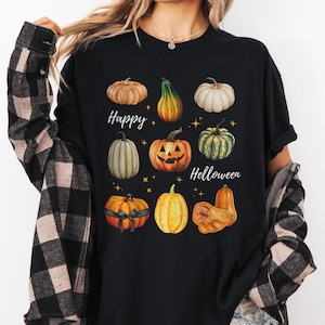 Può includere: T-shirt nera con illustrazioni ad acquerello di zucche nei toni dell'arancione, del verde e del bianco. Le parole "Happy Helloween" sono scritte in un carattere script, con piccole stelle dorate sparse intorno alle zucche.