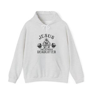 Peut inclure: Un sweat à capuche gris clair avec une capuche à cordon et une poche kangourou. Le devant présente un graphisme de Jésus soulevant des poids avec le texte "JESUS THE ULTIMATE DEADLIFTER" en noir.