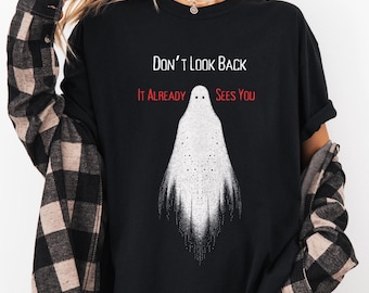 Op de afbeelding: Zwart T-shirt met de tekst "Don't Look Back, It Already Sees You" en een witte spookafbeelding. Het spook heeft een gestippeld lichaam en een gefranjerde onderkant. Het shirt is een casual stijl.