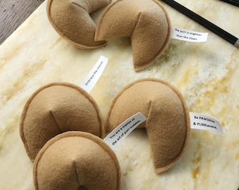 May include: A white paper takeout container filled with tan felt fortune cookies, accompanied by four loose felt fortune cookies and two black chopsticks. Each cookie contains a small white paper strip with printed text.