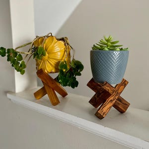 May include: Two decorative plant pots on wooden stands. One pot is yellow and shaped like a flower, holding a green plant. The other is gray with a textured pattern, holding a small succulent. The stands are made of crisscrossed wooden beams.