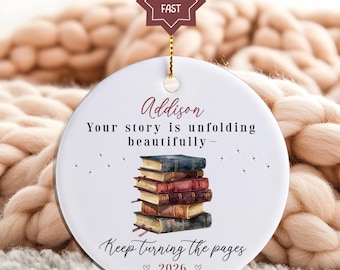 May include: A white ceramic ornament with a stack of books illustration. The ornament has text that reads "Addison, Your story is unfolding beautifully - Keep turning the pages 2026". A gold string and a star-shaped tag are attached.