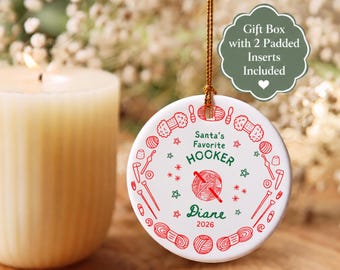 May include: A round white ceramic ornament hanging by a gold string. The design features red yarn balls and crochet hooks in a circular pattern. Text reads: Santa's Favorite HOOKER, Diane, and 2026. A green label states: Gift Box with 2 Padded Inserts Included.
