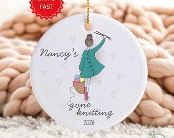 May include: White ceramic ornament with a gold string for hanging. The ornament features a cartoon illustration of a person in a green coat holding knitting needles and a basket of yarn. Text reads "Nancy's gone knitting 2026". Pastel hearts decorate the background.