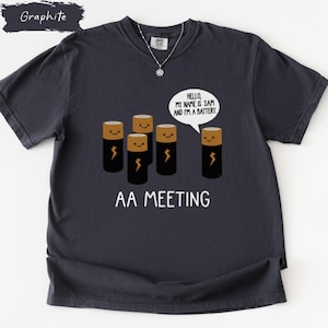 May include: Graphite-colored t-shirt with a humorous graphic. The design features five smiling battery cell characters with lightning bolt symbols, and the text "AA MEETING". A speech bubble says, "Hello, my name is Sam and I'm a battery."