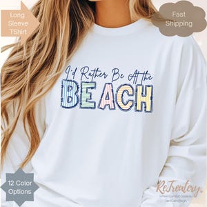 May include: White long-sleeve T-shirt with the phrase "I'd Rather Be At the BEACH" in colorful, striped letters. The shirt has a relaxed fit and is suitable for casual wear. The design includes a beach theme.
