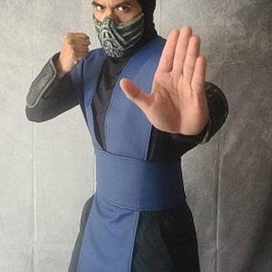 May include: A person in a blue and black ninja costume with a mask. The costume includes a blue tunic, belt, and arm guards. The person is making a hand gesture.