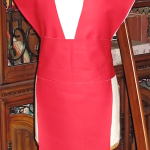 Puede incluir: Una prenda roja con cuello en V profundo y mangas anchas se exhibe en un maniquí. La prenda tiene un cinturón rojo a juego y una falda larga y recta. El maniquí está colocado contra un fondo de un armario de madera.