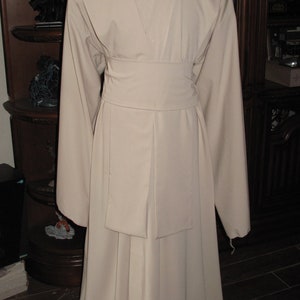 May include: A long, beige robe with a wide collar and a sash tied at the waist. The robe has long sleeves and a flowing skirt.