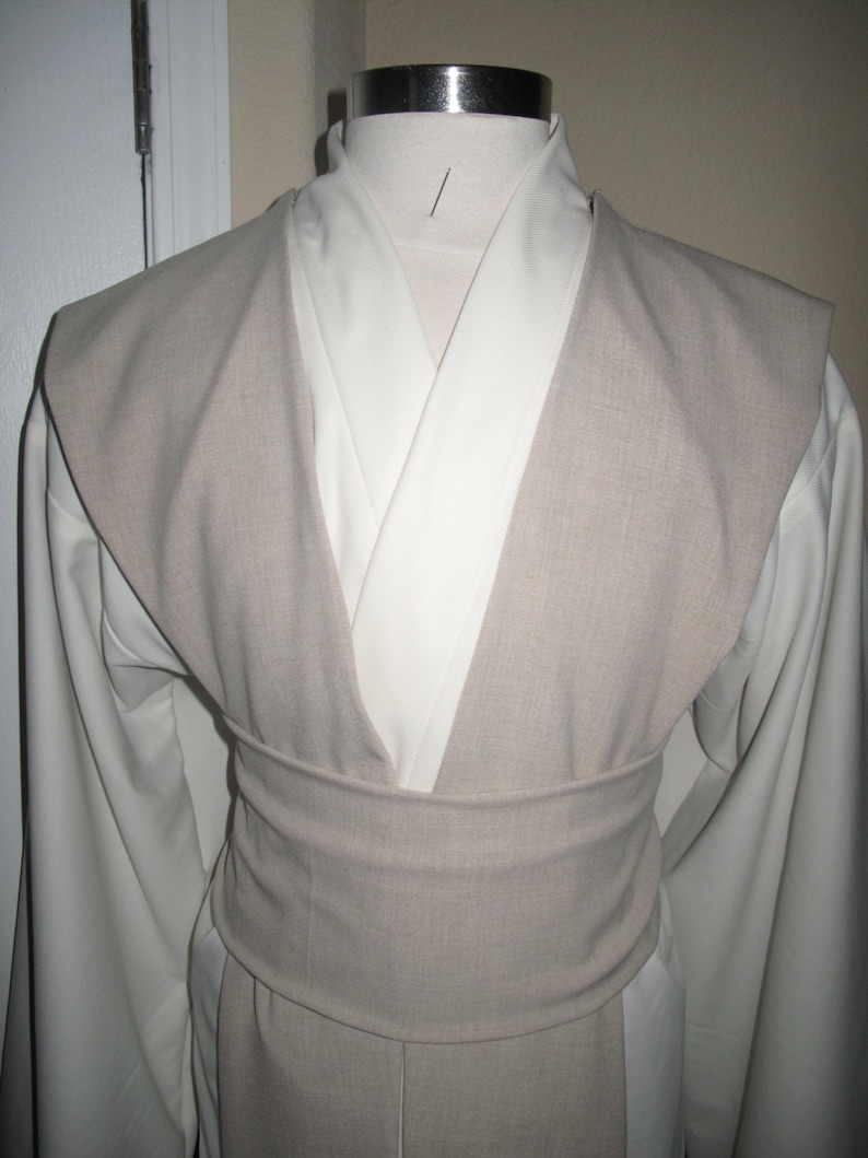 Pu&ograve; includere: Un costume beige chiaro e bianco esposto su un manichino. Il costume presenta un cappotto beige con un ampio colletto e una cintura abbinata. Lo strato inferiore &egrave; bianco, con maniche lunghe. Il costume &egrave; progettato con uno scollo a V.