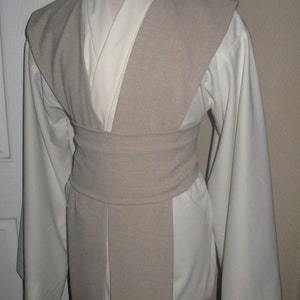 Pu&ograve; includere: Una tunica color crema con maniche lunghe e un tabarro e cintura beige. Il costume &egrave; esposto su un manichino, mostrando il design a strati e la vestibilit&agrave; su misura. L'outfit ricorda un Jedi o un personaggio simile.