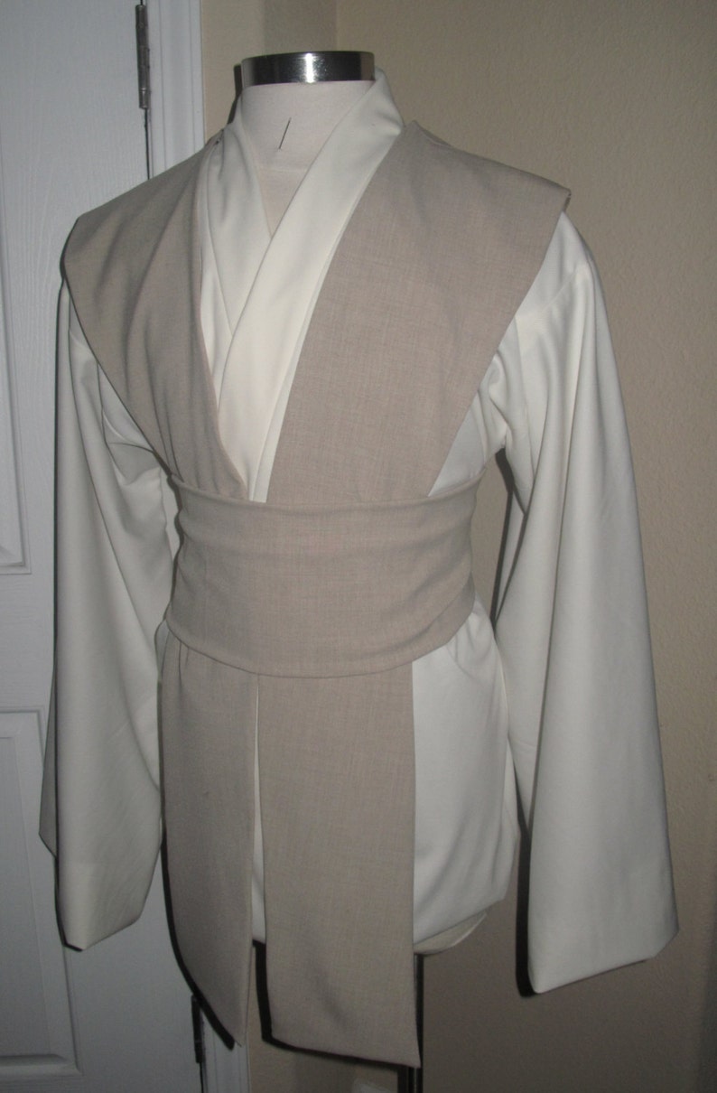 Pu&ograve; includere: Una tunica color crema con maniche lunghe e un tabarro e cintura beige. Il costume &egrave; esposto su un manichino, mostrando il design a strati e la vestibilit&agrave; su misura. L'outfit ricorda un Jedi o un personaggio simile.