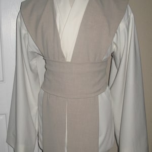 Pu&ograve; includere: Un completo tunica e tabarro Jedi color crema. La tunica ha maniche lunghe e ampie e un design a scollo a V. Un tabarro e una cintura color beige chiaro avvolgono la tunica. L'outfit &egrave; esposto su un manichino.