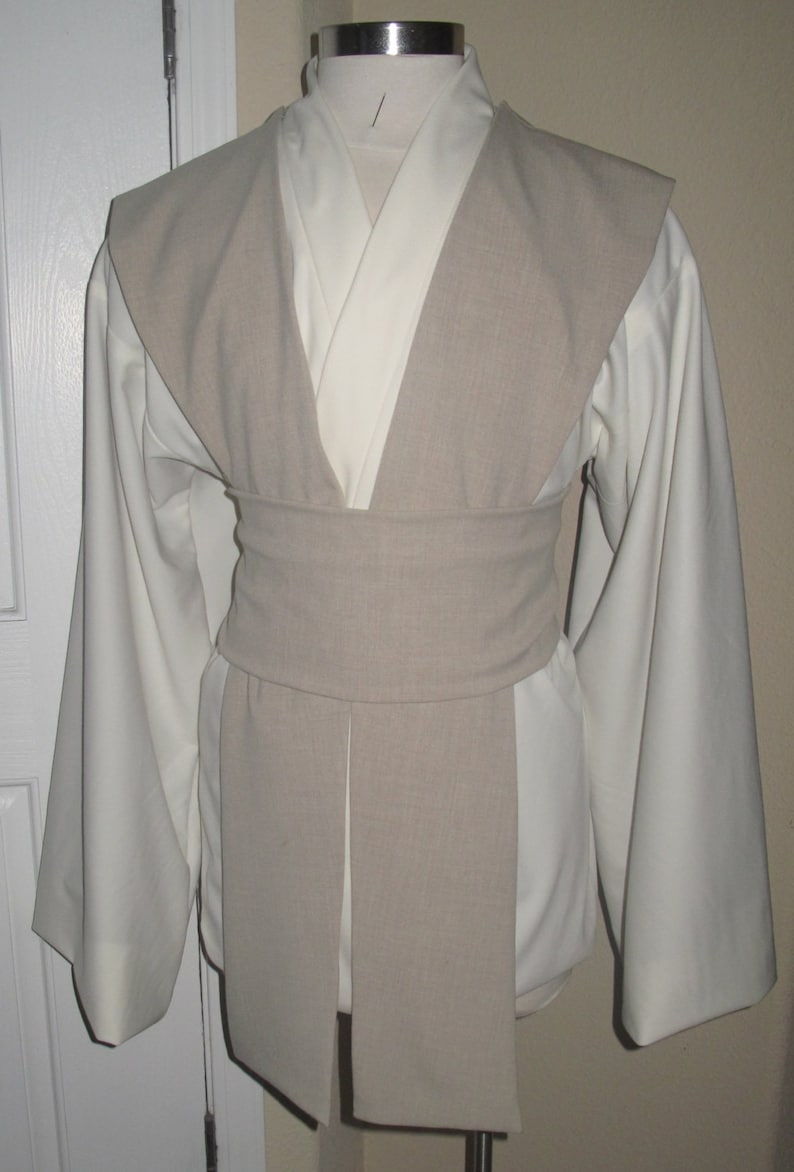 Pu&ograve; includere: Un completo tunica e tabarro Jedi color crema. La tunica ha maniche lunghe e ampie e un design a scollo a V. Un tabarro e una cintura color beige chiaro avvolgono la tunica. L'outfit &egrave; esposto su un manichino.