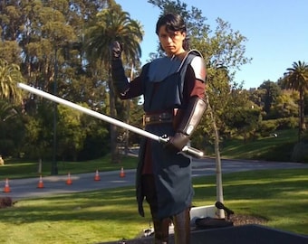 May include: A person dressed as a Jedi Knight, wearing a dark blue and brown costume, holds a white lightsaber in their right hand. They are standing on a black surface in front of a green grassy area.