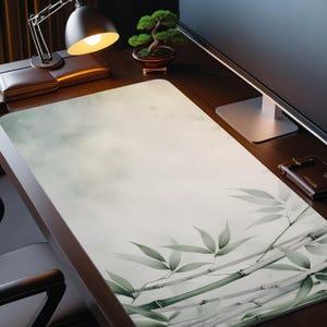 May include: A rectangular desk mat with a watercolor design of bamboo stalks and leaves in shades of green and gray. The background has a soft, cloudy appearance. The mat is on a dark wooden desk, with a lamp, bonsai tree, and monitor visible.