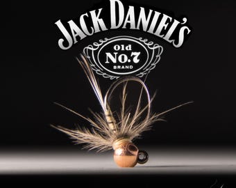 May include: A close-up of a fly fishing lure with a copper bead head and brown feathers. The lure is sitting on a white surface with a black background. The Jack Daniel's logo is visible in the background.