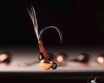 May include: A close-up of a copper bead fly fishing lure with a brown and orange thread body and white feathers.