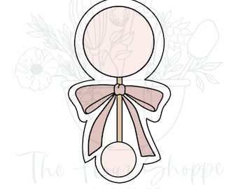 May include: A cookie cutter in the shape of a baby rattle. The rattle has a circular top and bottom, connected by a straight handle. A bow is tied around the handle. The design is outlined in black, with a light pink color fill.