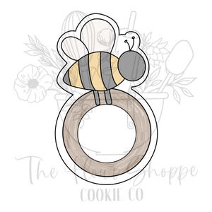 May include: A bee-shaped cookie cutter with a yellow and gray striped body, gray head, and a wooden ring base. The cutter has a black outline and is designed for baking. The text "The Flour Shoppe Cookie Co" is at the bottom.