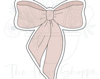 May include: A light pink bow-shaped cookie cutter with a black outline. The bow has two long, flowing tails and a detailed center knot. The background features a floral design and the text "The Flour Shoppe Cookie Co."