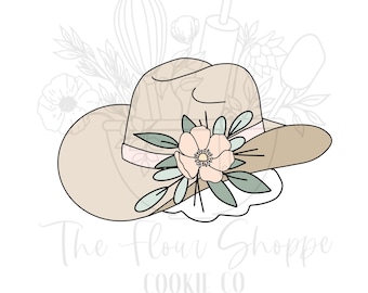 May include: A tan cowboy hat cookie cutter with a pink ribbon and a floral arrangement. The hat has a wide brim and is decorated with a light pink flower and green leaves. The text "The Flour Shoppe Cookie Co" is at the bottom.