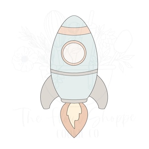 May include: A light blue rocket ship cookie cutter with a round window and peach accents. The rocket has gray fins and a peach-colored flame. The text "The Flour Shoppe Cookie Co" is at the bottom.