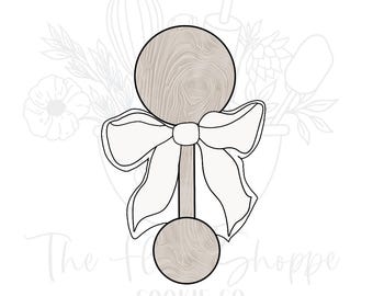 May include: A wooden rattle with a white bow. The rattle is made of two round pieces of wood connected by a stick. The bow is tied around the stick.