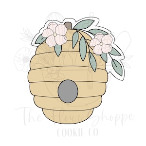 May include: A light tan beehive cookie cutter with a gray entrance and a floral design. The flowers are light pink with green leaves. The text "The Flour Shoppe Cookie Co" is at the bottom.