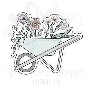 May include: A cookie cutter in the shape of a wheelbarrow filled with flowers. The wheelbarrow is light blue with a gray wheel and handle. The flowers are pink, purple, and white with green leaves. The Flour Shoppe Cookie Co. is written at the bottom.