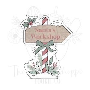 May include: A cookie cutter in the shape of a signpost pointing right, with the text "Santa's Workshop". The signpost is decorated with a green bow, holly, and winter foliage. The pole has red and white stripes.