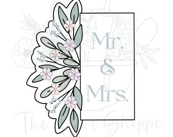 May include: A cookie cutter with a floral design and the words "Mr. & Mrs." in a rectangular frame. The floral design includes green leaves and small pink and white flowers.