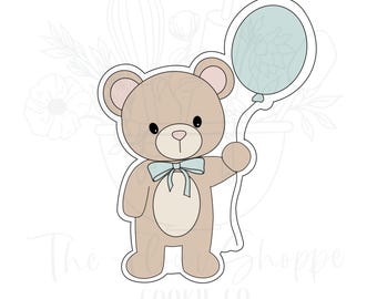 May include: A cookie cutter in the shape of a teddy bear holding a balloon. The bear is light brown with a cream-colored belly and a light blue bow tie. The balloon is light blue and has a floral design inside.