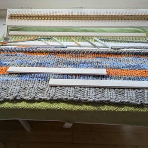 Puede incluir: Un telar de punto con una tela tejida gris, azul y naranja sobre él. Hay tres agujas de tejer de madera blanca encima de la tela.