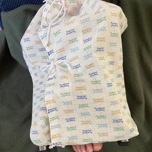 Puede incluir: Una bolsa de tela blanca con un patrón repetido del texto "Hospital Property" en azul, verde y dorado. La bolsa está hecha de una tela suave y natural y tiene un cierre de cordón.