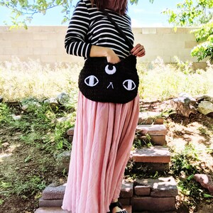 May include: A black crocheted cat-shaped shoulder bag with white eyes and a crescent moon on the forehead. The bag is worn by a person wearing a striped shirt and a pink skirt.